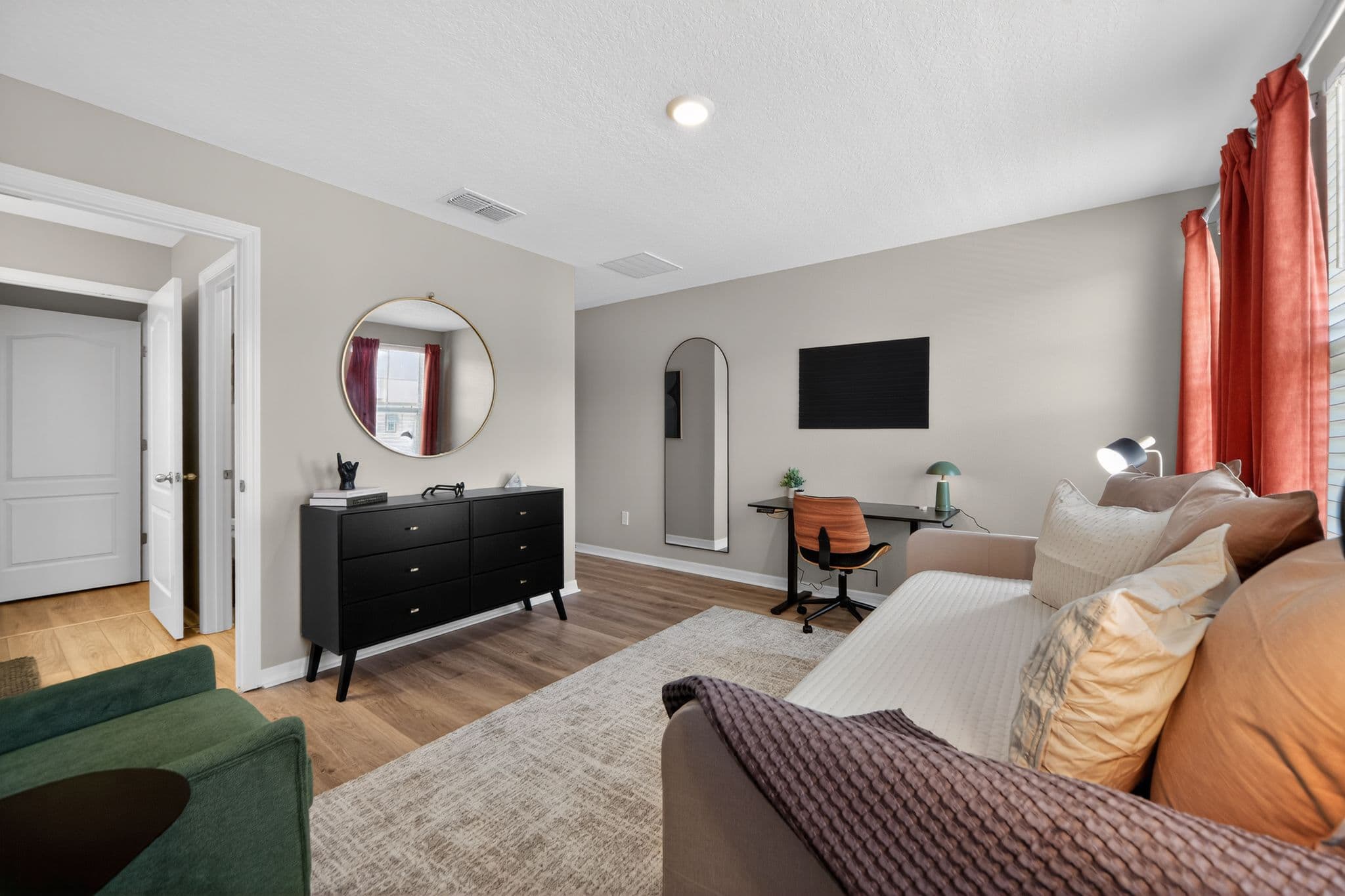 Modern bedroom featuring a black dresser, round mirror, desk, and daybed with orange accents.