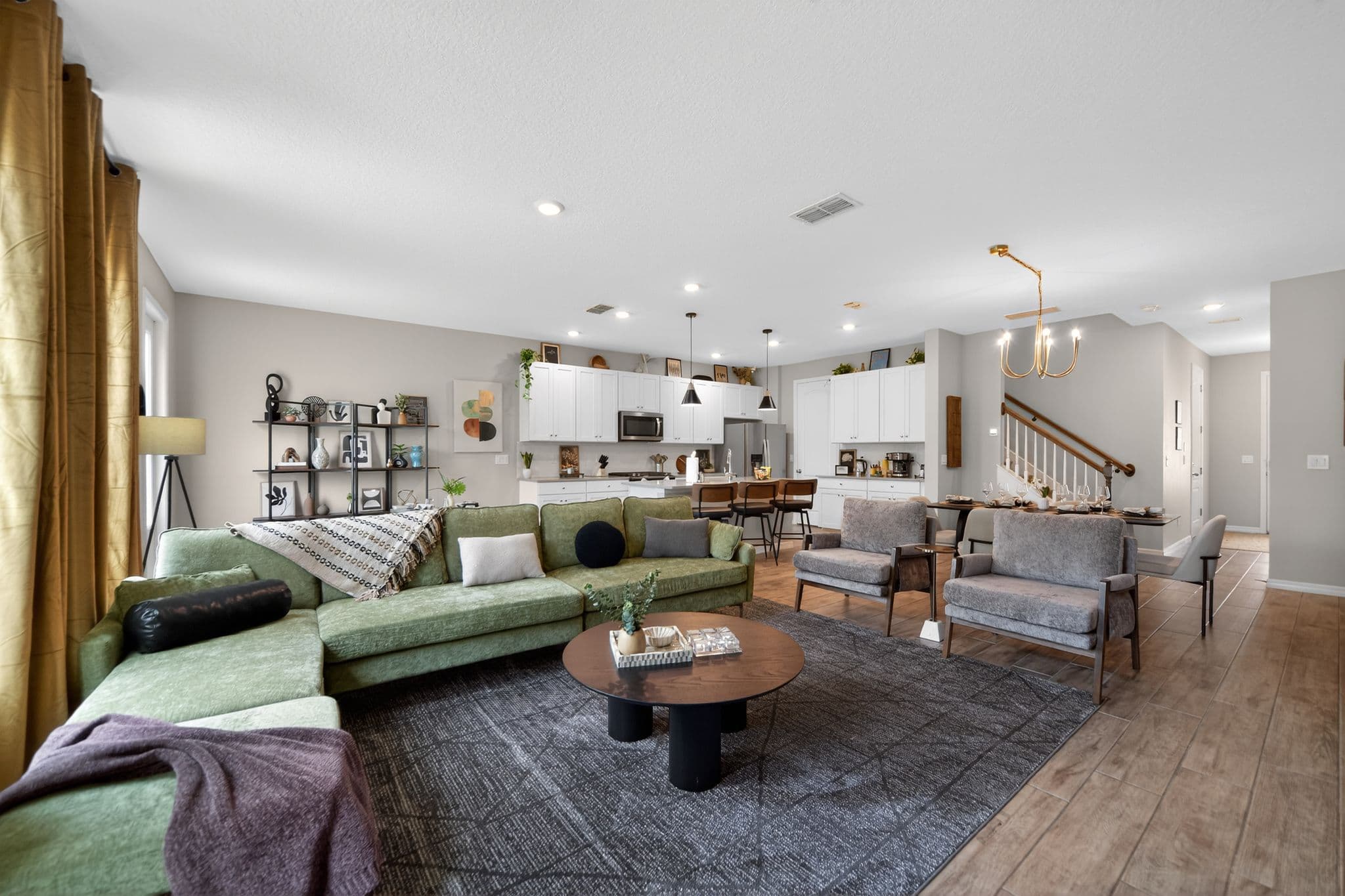 Modern open-concept living area with a green sectional sofa, grey armchairs, and white kitchen.
