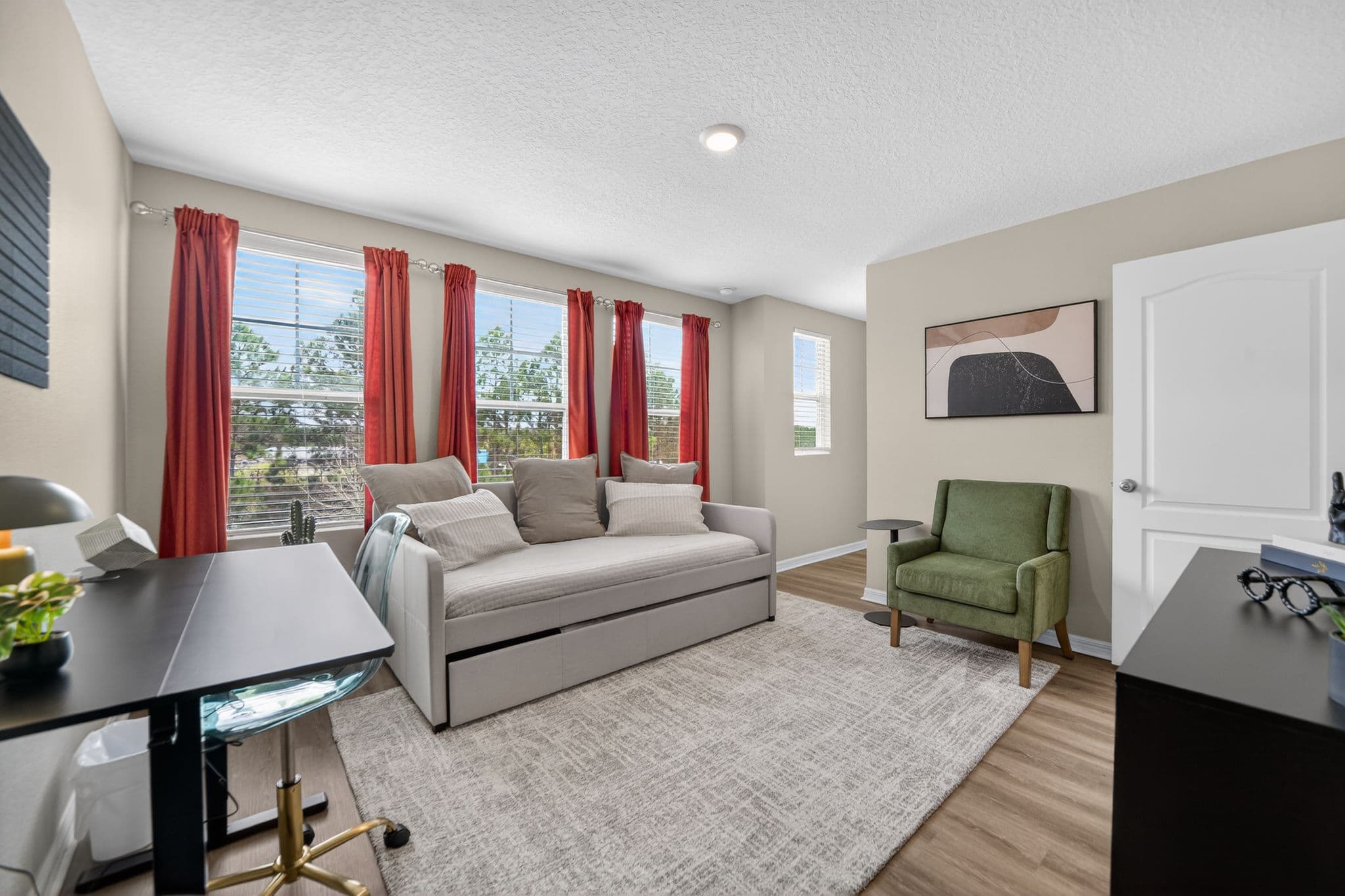 Modern home office featuring a gray daybed, red curtains, green armchair, and black desk.