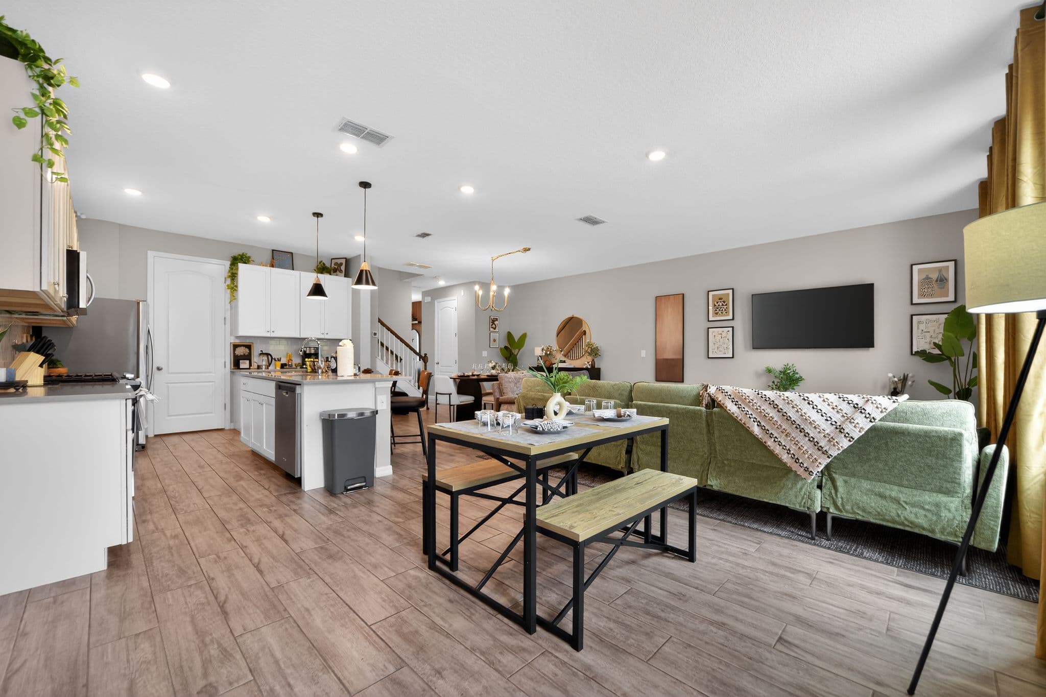 Modern open-concept living space with white kitchen, wooden dining table, and green sectional sofa.