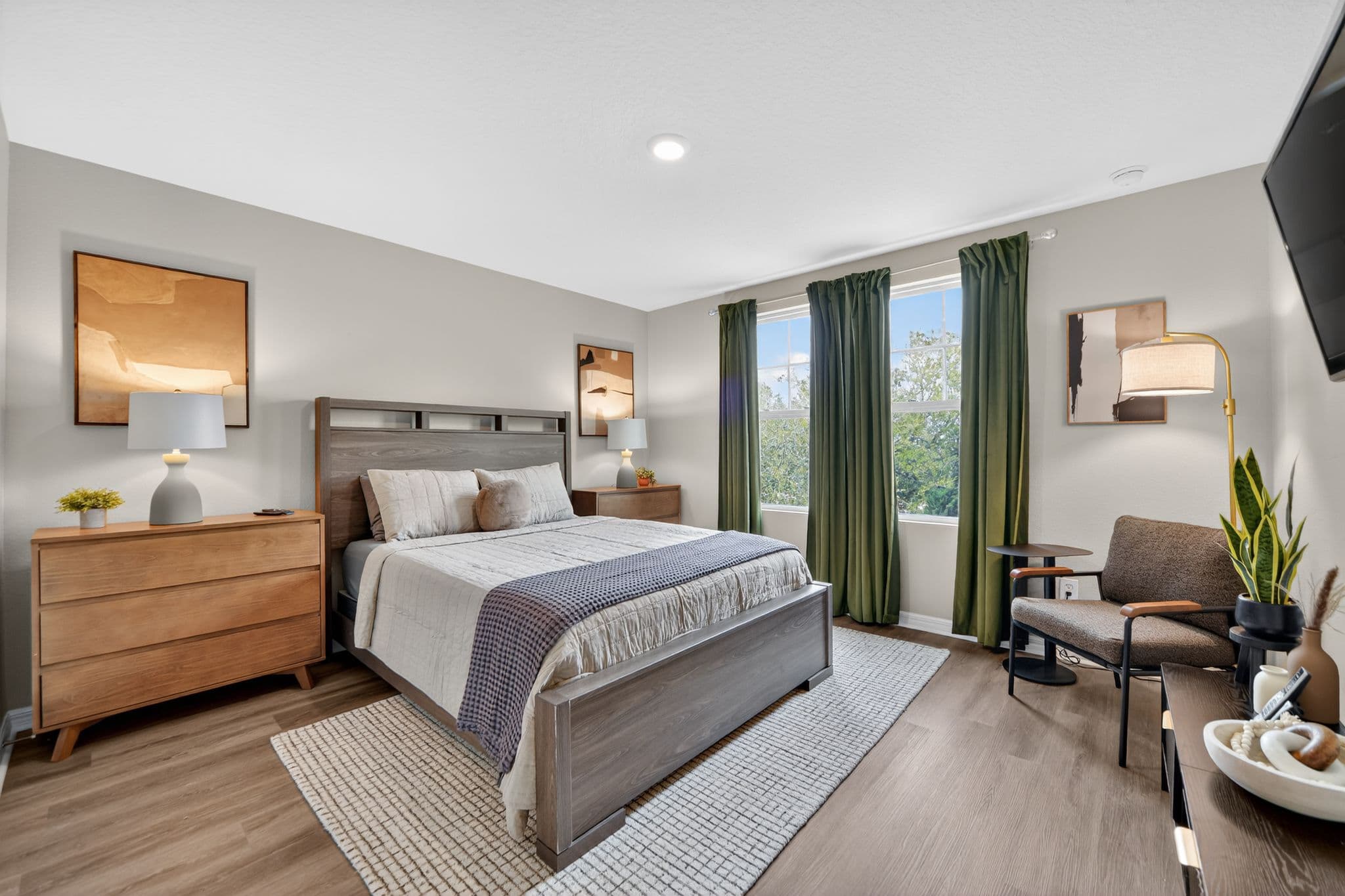 Modern bedroom featuring a wooden bed, olive green curtains, grey armchair, and neutral decor.