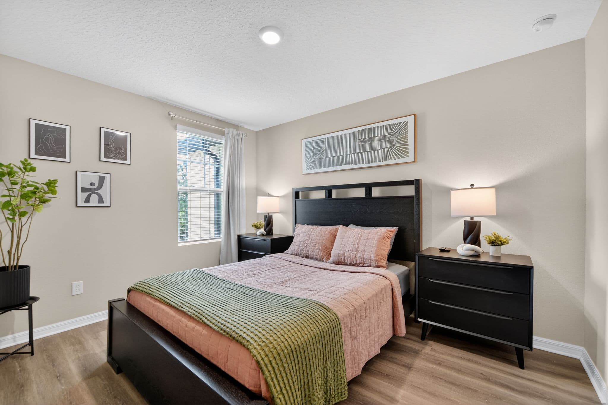 Modern bedroom with black bed, pink bedding, green throw, matching nightstands, and abstract wall art.