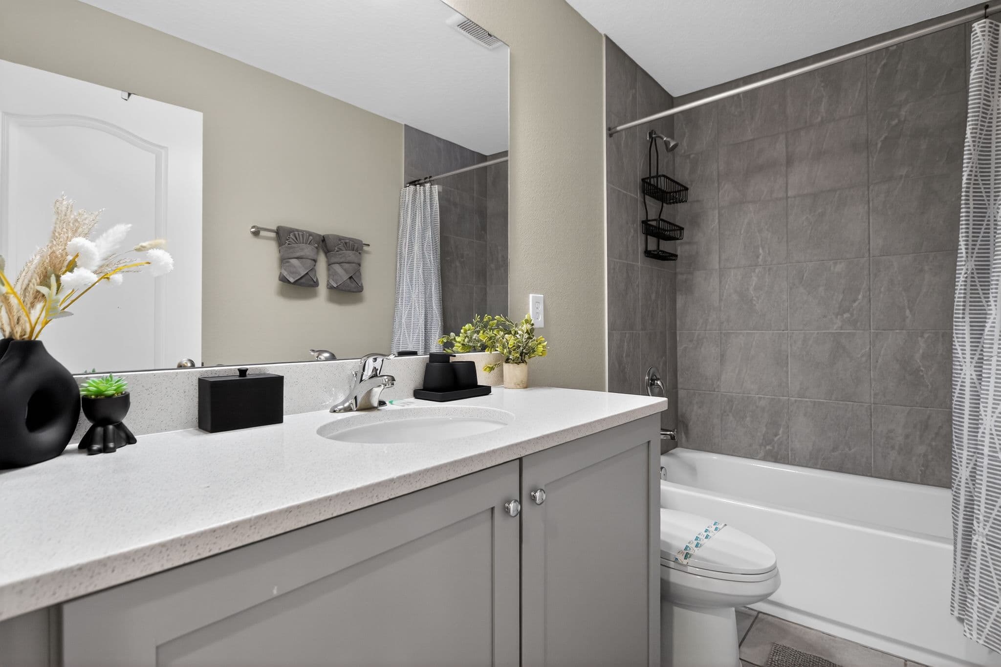 Modern bathroom with grey vanity, white countertop, large mirror, and grey tiled shower area.